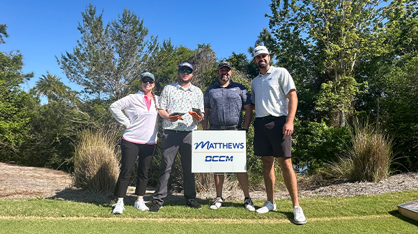Smiling group of golfers standing behind a sign with a Matthews DCCM logo on it